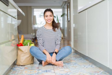 Yüzünde mutlu ve güzel bir gülümseme ile mutfakta taze yiyecek dolu kese kağıdı ile oturan bir genç bayan. Şanslı kişi.