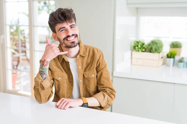 Young man wearing casual jacket sitting on white table smiling doing phone gesture with hand and fingers like talking on the telephone. Communicating concepts.