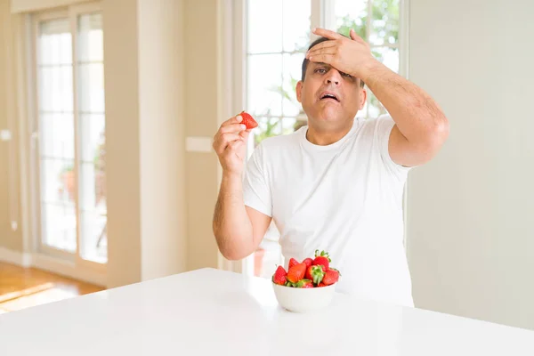 中年男子在家里吃草莓 用手在头上强调 羞愧和惊讶的脸震惊 愤怒和沮丧 因错误而恐惧和不安 — 图库照片