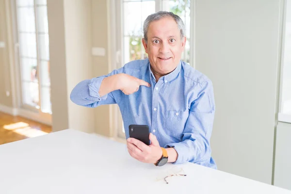 Handsome senior man using smartphone sending message with surprise face pointing finger to himself