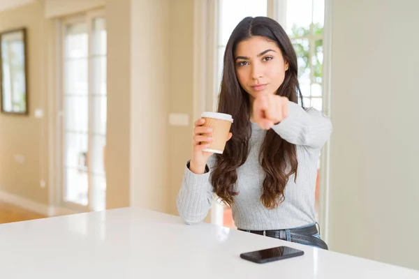 Young woman drinking a cup of coffee at home pointing with finger to the camera and to you, hand sign, positive and confident gesture from the front