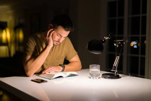 Joven hombre guapo estudiando en casa, leyendo un libro por la noche — Foto de Stock