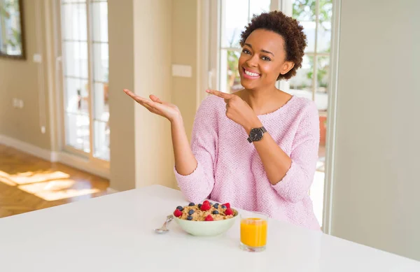 年轻的非洲裔美国妇女在早上在家里吃健康的早餐惊讶和微笑着对着镜头 同时提出用手和手指指指点点 — 图库照片