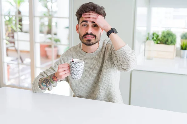 Young man drinking a cup of coffee in the morning stressed with hand on head, shocked with shame and surprise face, angry and frustrated. Fear and upset for mistake.
