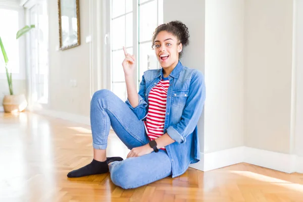 Beautiful young african american woman with afro hair sitting on the floor with a big smile on face, pointing with hand and finger to the side looking at the camera.