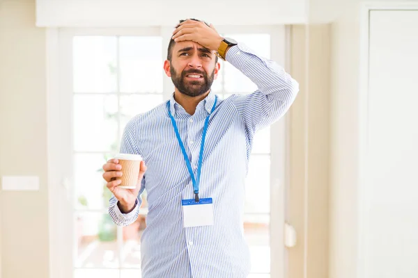 Handsome hispanic man wearing id card and drinking a cup of coffee stressed with hand on head, shocked with shame and surprise face, angry and frustrated. Fear and upset for mistake.