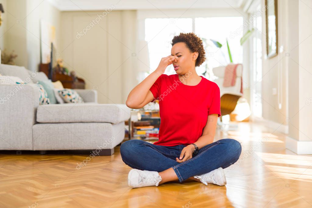 Young beautiful african american woman sitting on the floor at home smelling something stinky and disgusting, intolerable smell, holding breath with fingers on nose. Bad smells concept.