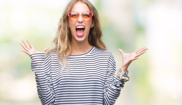 Beautiful young blonde woman wearing sunglasses over isolated background crazy and mad shouting and yelling with aggressive expression and arms raised. Frustration concept.