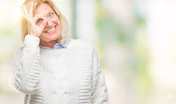 Middle age blonde woman wearing winter sweater over isolated background doing ok gesture with hand smiling, eye looking through fingers with happy face.