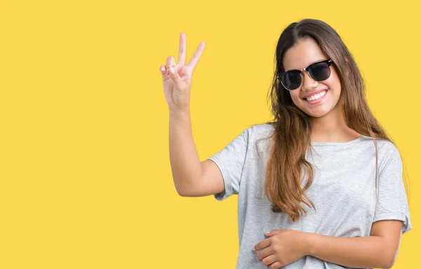 Young beautiful brunette woman wearing sunglasses over isolated background smiling with happy face winking at the camera doing victory sign. Number two.