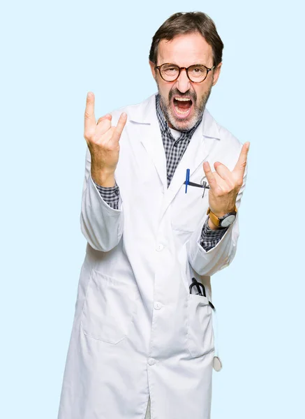 Middle age doctor men wearing medical coat shouting with crazy expression doing rock symbol with hands up. Music star. Heavy concept.