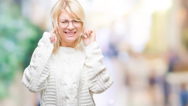 若い美しい金髪女性の騒々しい音楽のノイズの腹式指で耳を覆う孤立した背景に冬のセーターとメガネを着用します 聴覚障害者の概念 — ストック写真