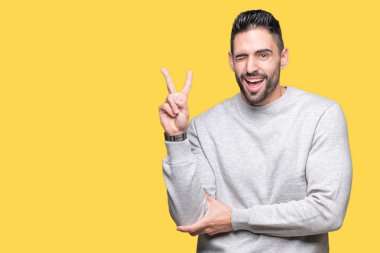 Mutlu yüz zafer işareti yaparken kameraya göz kırpıyor ile gülümseyen izole arka plan üzerinde sweatshirt giyen genç yakışıklı adam. İki numaralı.