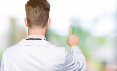 Tıbbi giyen genç yakışıklı doktor adam kat geriye doğru arkasında parmak eliyle işaret Posing
