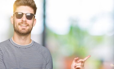 Kameraya bakarak yan el ve parmak ile işaret eden genç yakışıklı adam, yüzünde kocaman bir gülümseme ile güneş gözlüğü takıyor.