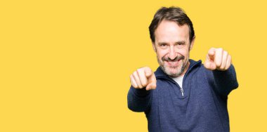 Orta yaş yakışıklı adam kazak giyiyor ve kameranın olumlu ve neşeli gülümseyen parmaklarıyla işaret