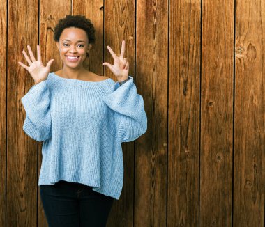 Gösterilen ve parmakları ile işaret izole arka plan üzerinde bir kazak giyen genç güzel Afrikalı-Amerikalı kadın kendinden emin ve mutlu gülümseyerek süre sekiz numara.