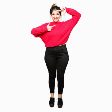 Kırmızı kazak ve elle yapma çerçeve ve parmakları ile mutlu yüz gülümseyen topuz giyen genç güzel kadın. Yaratıcılık ve Fotoğrafçılık kavramı.