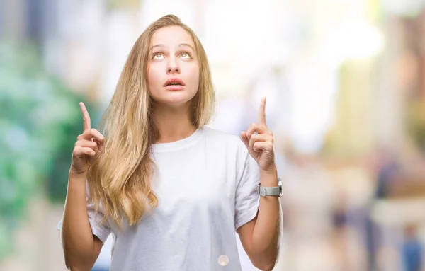 年轻美丽的金发碧眼的女人穿着休闲白色 T恤在孤立的背景惊讶和惊讶地抬头 用手指指指点点 举起双臂 — 图库照片