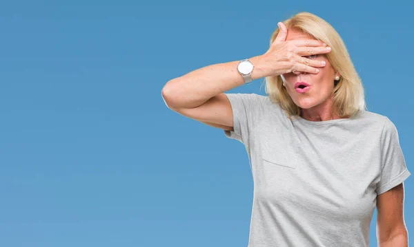 Middle age blonde woman over isolated background peeking in shock covering face and eyes with hand, looking through fingers with embarrassed expression.