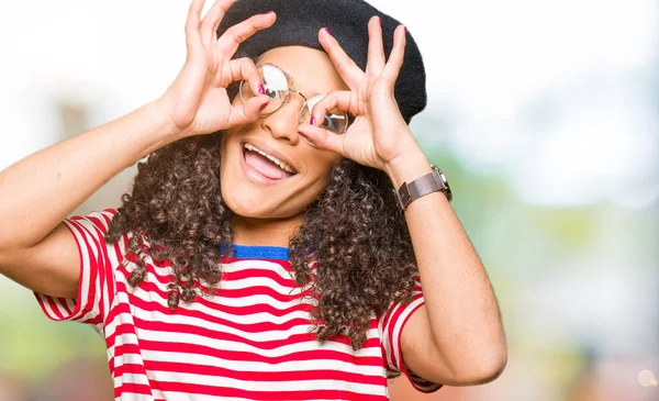 Young beautiful woman with curly hair wearing glasses and fashion beret doing ok gesture like binoculars sticking tongue out, eyes looking through fingers. Crazy expression.