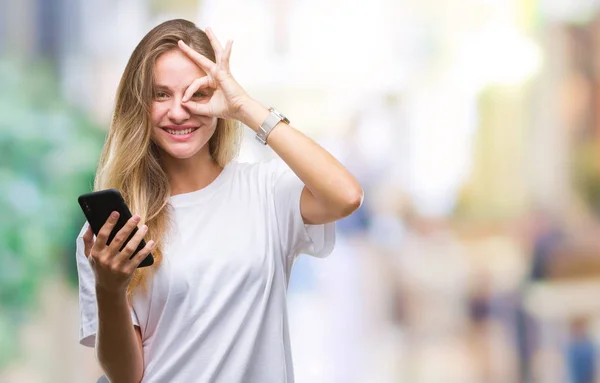 年轻漂亮的金发女人使用智能手机在孤立的背景下 快乐的脸微笑做 签名与手在眼睛看通过手指 — 图库照片