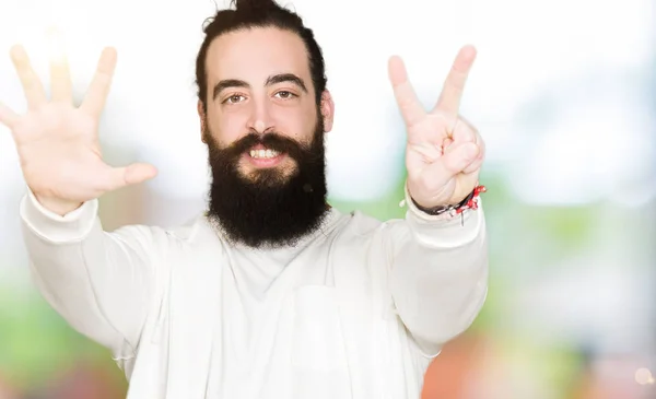 Young man with long hair and beard wearing sporty sweatshirt showing and pointing up with fingers number seven while smiling confident and happy.