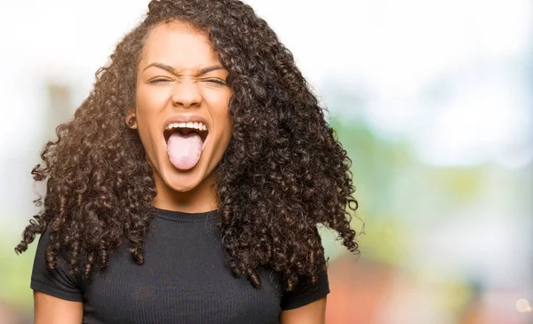 Young beautiful woman with curly hair sticking tongue out happy with funny expression. Emotion concept.