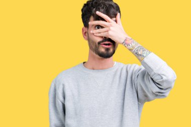 Mahcup ifadeyle parmaklarımın arasından seyir yüz ve gözler el ile kapsayan şok bakmak izole arka plan üzerinde sweatshirt giyen genç yakışıklı adam.