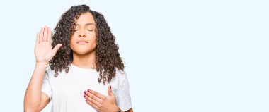 Kıvırcık saçlı bir sadakat sözü yemin yapma beyaz t-shirt Swearing göğüs ve avuç, el ile giyen genç güzel kadın