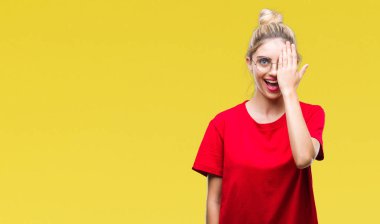 El yüz ve sürpriz duygu kendinden emin gülümseme ile tek gözüyle kapsayan izole arka plan üzerinde kırmızı t-shirt ve gözlük giyen genç güzel sarışın kadın.