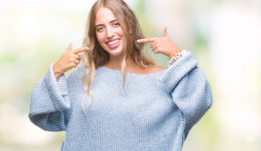 Kendinden emin gösterilen ve işaret parmakları diş ve ağız ile gülümseyen izole arka plan üzerinde kış kazak giyen güzel genç sarışın kadın. Sağlık kavramı.