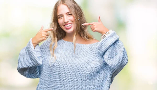 Kendinden emin gösterilen ve işaret parmakları diş ve ağız ile gülümseyen izole arka plan üzerinde kış kazak giyen güzel genç sarışın kadın. Sağlık kavramı.