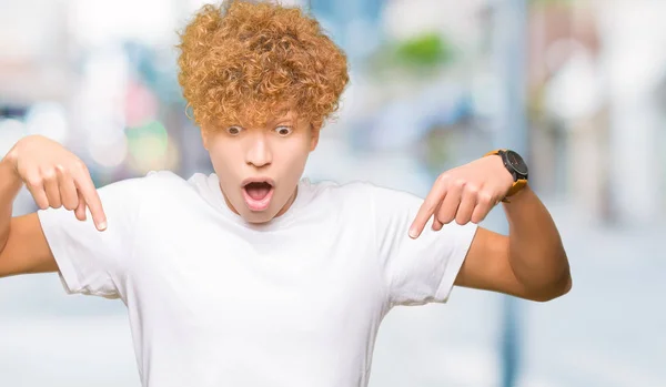 Young handsome man with afro hair wearing casual white t-shirt Pointing down with fingers showing advertisement, surprised face and open mouth