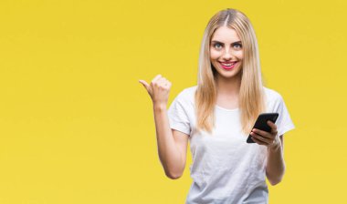 Genç güzel sarışın kadın işaret ve başparmak kadar mutlu yüz gülümseyen tarafı ile gösteren izole arka plan üzerinde akıllı telefon kullanma