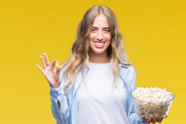 Güzel genç sarışın kadın ayakta ve diş gösterilen kendine güvenen bir gülümsemeyle gülümseyerek mutlu bir yüz ile izole arka plan üzerinde patlamış mısır yemek