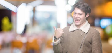 Yakışıklı delikanlı arıyor ve başparmak yanında yukarıyı mutlu yüz ile gülümseyen izole arka plan üzerinde kış palto giyiyor.