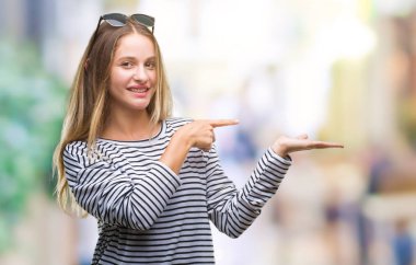 Genç güzel sarışın kadın hayrete ve el ile sunulması ve parmak ile işaret ederken kameraya gülümseyen izole arka plan üzerinde güneş gözlüğü takıyor.