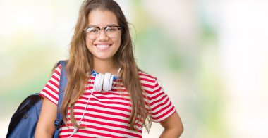 Yüzünde mutlu ve güzel bir gülümseme ile izole arka plan üzerinde kulaklık ve sırt çantası giyen genç güzel esmer öğrenci kadın. Şanslı kişi.