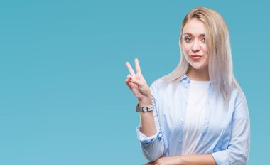 Genç Sarışın kadın mutlu yüz zafer işareti yaparken kameraya göz kırpıyor ile gülümseyen izole arka plan yüzünden. İki numaralı.