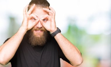 Dilini, parmaklarının arasından baktığını yapışmasını dürbün gibi ok jest yapıyor rahat t-shirt giyen genç sarışın adam. Deli ifade.