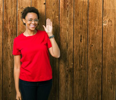 İzole arka plan Waiving mutlu selam söyleyerek üzerinde gözlük ve gülümseyen, dostça karşılama jest giyen güzel genç Afro-Amerikan kadın