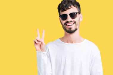 Genç yakışıklı adam mutlu yüz zafer işareti yaparken kameraya göz kırpıyor ile gülümseyen izole arka plan üzerinde güneş gözlüğü takıyor. İki numaralı.