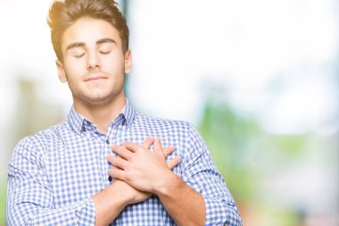 Genç yakışıklı iş adamı izole arka plan ile gülümseyen üzerinde kapalı gözler ve yüzünde minnettar jest ile göğüs üzerinde eller. Sağlık kavramı.