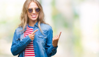 Güzel genç sarışın kadın gülümseyerek ve iki elleri ve parmakları ile tarafına işaret kamera bakarak izole arka plan üzerinde güneş gözlüğü takıyor.