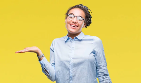 Young braided hair african american business girl wearing glasses over isolated background smiling cheerful presenting and pointing with palm of hand looking at the camera.