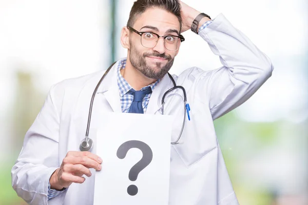 Handsome young doctor man holding paper with question mark over isolated background stressed with hand on head, shocked with shame and surprise face, angry and frustrated. Fear and upset for mistake.