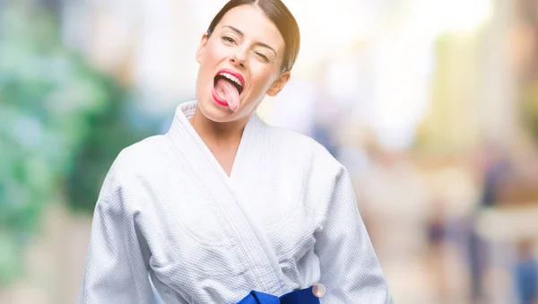 Young beautiful woman wearing karate kimono uniform over isolated background sticking tongue out happy with funny expression. Emotion concept.