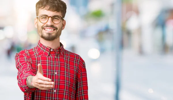 Young handsome man wearing glasses over isolated background smiling friendly offering handshake as greeting and welcoming. Successful business.