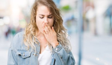 İzole arka plan üzerinde kot ceket giyen güzel genç sarışın kadın el ağzıyla kapsayan yorgun esneme sıkılmış. Restless ve uyku hali.